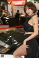 A woman in a black dress posing next to a car.