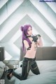 A woman with purple hair sitting on a bed holding a gun.