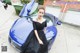 A woman in a black dress standing next to a blue car.