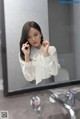 A woman in a white shirt is brushing her teeth in a bathroom mirror.