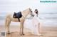 A woman in a white dress standing next to a horse on the beach.