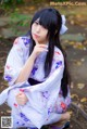 A woman in a kimono sitting on the ground with her hand on her chin.