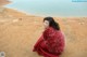 A woman wrapped in a red blanket sitting on a rock by the water.