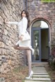 A woman leaning against a stone wall in a white dress.