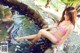 A woman in a red and white bikini sitting on a rock by a pond.