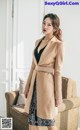 A woman standing in front of a couch wearing a beige coat.